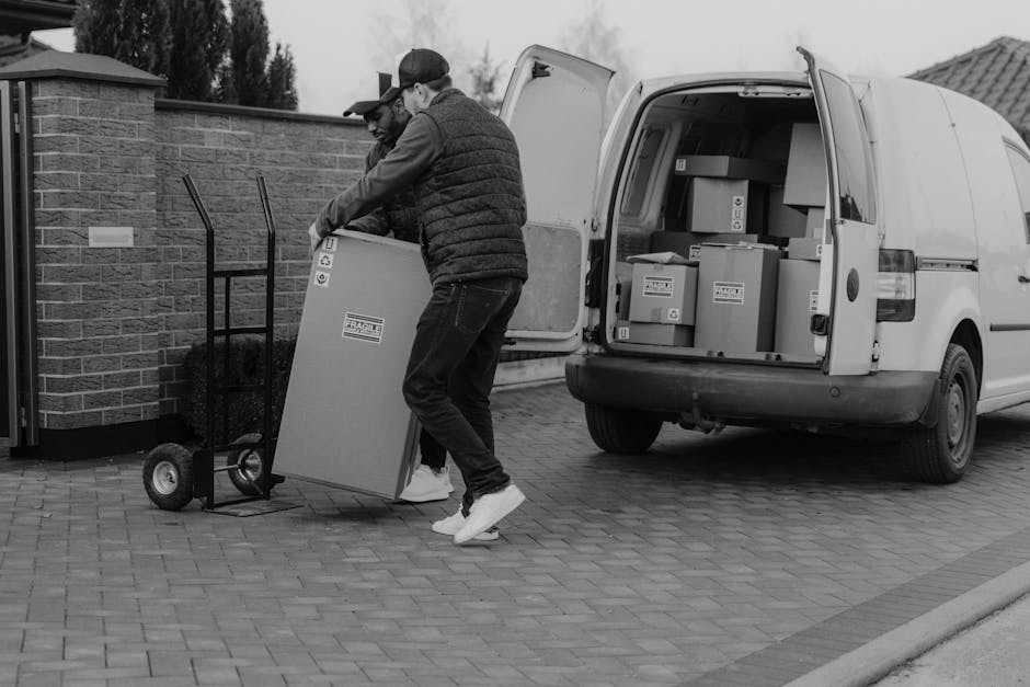 Two men are engaged in a home relocation process outdoors, with a white van parked on a brick driveway and the van's rear doors open, revealing packed boxes inside. One man, wearing a black vest, grey