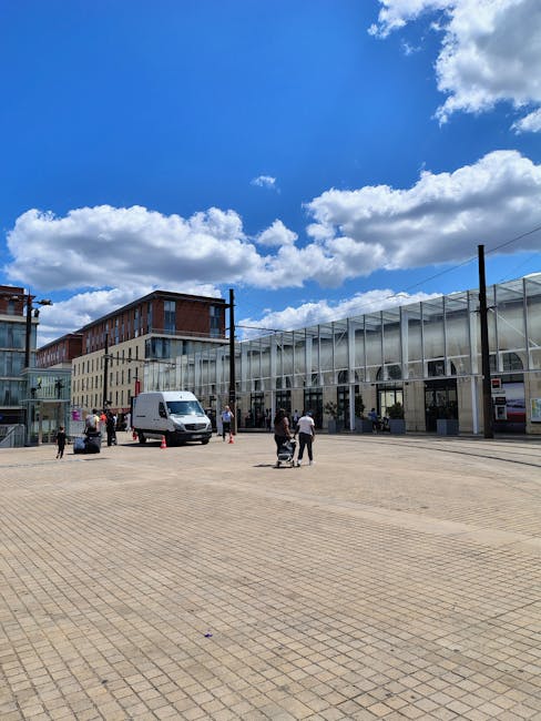 A clear outdoor area with a large paved open space adjacent to a modern glass-fronted building, possibly a station or commercial complex, under a bright blue sky with scattered white clouds. Several people are present, some pushing carts or walking, and a white delivery van is parked near the building. In the background, there are multi-storey buildings with a mix of brick and glass facades, indicating an urban environment. The scene suggests an active setting where items such as boxes, furniture, or appliances could be being loaded or unloaded, consistent with ground-level home relocation or furniture transport activities supported by companies like Man with Van Bow.