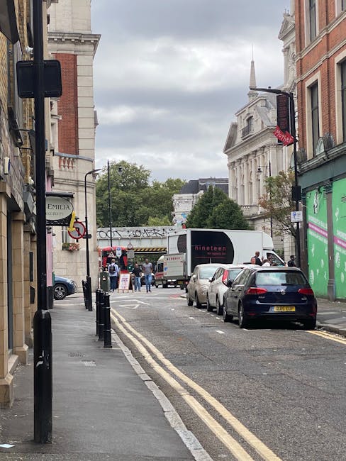 A narrow city street with parked cars lining the kerbside and a delivery van positioned in the middle of the road, partially blocking the lane. The sidewalk on the left side features metal bollards, a shop sign, and a storefront with a window display. Pedestrians are visible further down the street, some entering or leaving buildings, while others walk along the pavement. In the background, a bridge with decorative railings spans the street, and historical buildings with classical architecture, including a prominent white building with columns and a spire, are visible under an overcast sky. The scene depicts a typical urban environment suitable for house removals, with potential loading and unloading areas, and is consistent with an area serviced by professional removal companies such as Man with Van Bow, operating near Bow Road station.